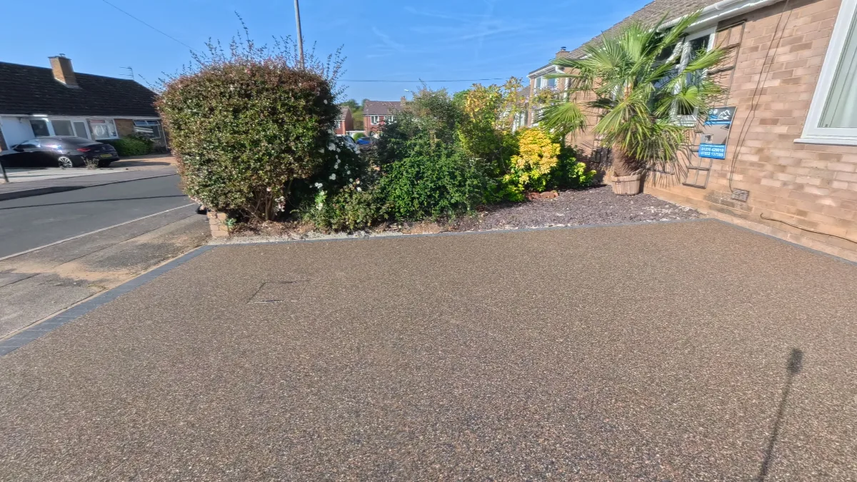 A completed resin bound driveway in a warm gravel tone, edged with charcoal block paving, running from the road up to a brick bungalow with a planted border.